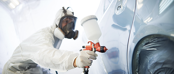 Worker wearing protective gear spray-painting a car in an auto body shop.
