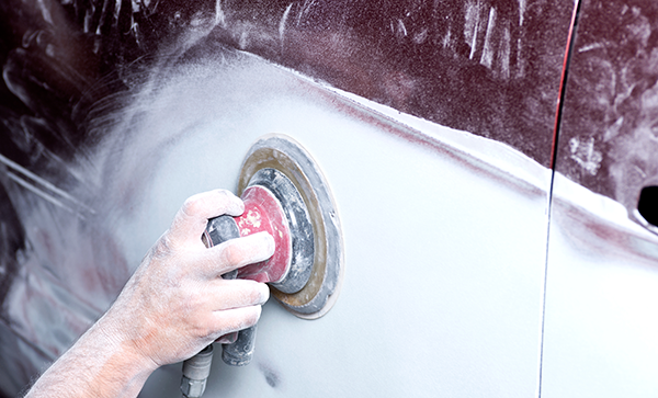 Close-up of a person&rsquo;s hand using an electric sander to smooth the surface of a car door during auto body repair. The car is partially sanded, with white dust covering the area.